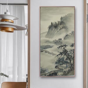 Könnte beinhalten: Gerahmtes Landschaftsgemälde in gedämpften Grau- und Beigetönen. Das Kunstwerk zeigt eine neblige Berglandschaft mit Bäumen, einem kleinen Gebäude und einer Brücke. Das Gemälde wird in einem Raum mit Holzmöbeln ausgestellt.