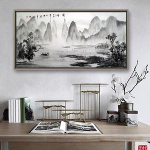 K&ouml;nnte beinhalten: Ein gerahmtes Schwarzwei&szlig;-Tuschbild eines Berglands mit einem Boot im Wasser. Das Bild h&auml;ngt an einer wei&szlig;en Wand &uuml;ber einem Holztisch mit einem Buch, dekorativen Metallst&auml;ndern und einer Vase mit wei&szlig;en Blumen.