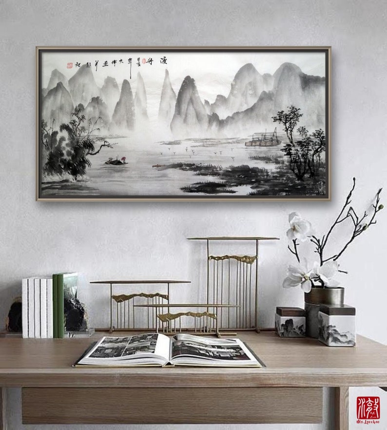 K&ouml;nnte beinhalten: Ein gerahmtes Schwarzwei&szlig;-Tuschbild eines Berglands mit einem Boot im Wasser. Das Bild h&auml;ngt an einer wei&szlig;en Wand &uuml;ber einem Holztisch mit einem Buch, dekorativen Metallst&auml;ndern und einer Vase mit wei&szlig;en Blumen.