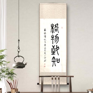 Könnte beinhalten: Eine traditionelle chinesische Schriftrolle mit schwarzer Kalligraphie hängt über einem hölzernen Teetisch. Auf dem Tisch stehen eine Teekanne, Tassen und Teezubehör. Eine hängende Teekanne aus Metall und Grünzeug tragen zur ruhigen Atmosphäre bei.