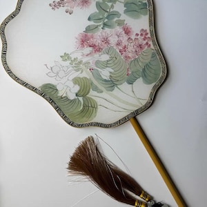 May include: A decorative hand fan with a floral design. The fan has a white background with pink flowers and green leaves. A brown tassel and a wooden handle are attached to the fan.