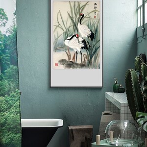 Puede incluir: Una pintura al agua enmarcada de dos grullas blancas de pie en un pantano con juncos verdes. La pintura tiene un fondo blanco y un marco negro.