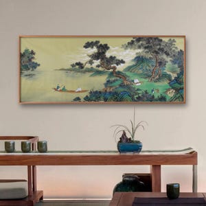 Könnte beinhalten: Gerahmtes Landschaftsgemälde, das eine ruhige Szene mit einem Boot auf einem See, Bäumen und Bergen darstellt. Das Kunstwerk weist gedämpfte Farben und einen traditionellen asiatischen Kunststil auf. Der Rahmen ist hellbraun.