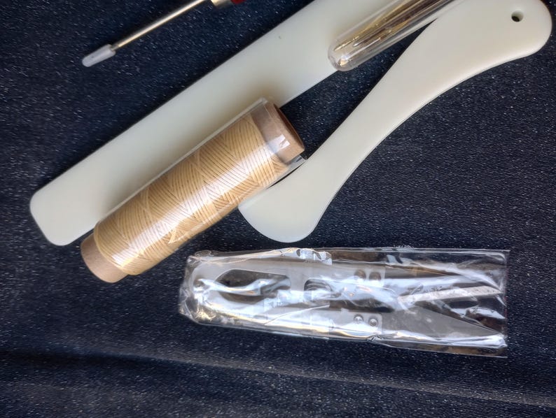 Peut inclure: Un ensemble d'outils de couture comprenant une bobine de fil beige, une petite paire de ciseaux et un outil en plastique blanc avec une aiguille.