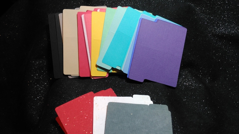 May include: A selection of colourful, rectangular cardstock tabs in various shades, including black, beige, pink, yellow, teal, and purple. The tabs have a notched design on one side. Some tabs have a textured finish, others a smooth surface.