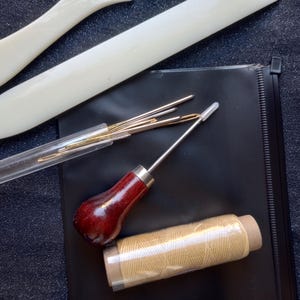 Peut inclure: Un kit de maroquinerie avec des outils comprenant un poin&ccedil;on &agrave; coudre, des aiguilles, du fil et un os &agrave; plier. Les outils sont dispos&eacute;s sur une surface noire.