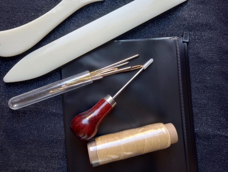 Peut inclure: Un kit de maroquinerie avec des outils comprenant un poin&ccedil;on &agrave; coudre, des aiguilles, du fil et un os &agrave; plier. Les outils sont dispos&eacute;s sur une surface noire.