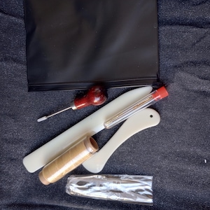May include: A leatherworking kit with tools and supplies. The kit includes a leather awl, a stitching pony, a tube of needles, a spool of waxed thread, and a black zippered pouch.