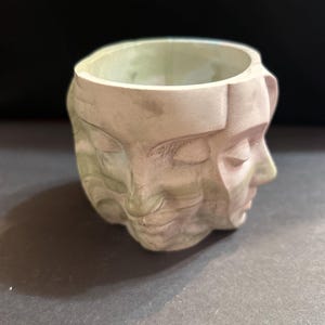 May include: A light beige ceramic planter with a unique design featuring multiple faces. The faces are sculpted with closed eyes and serene expressions. The planter has a round opening and is suitable for small plants or decorative use.
