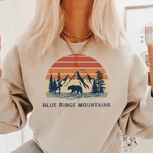 May include: A beige sweatshirt with a vintage-style graphic of mountains, a bear, and a sunset. The text "BLUE RIDGE MOUNTAINS" is printed below the graphic.