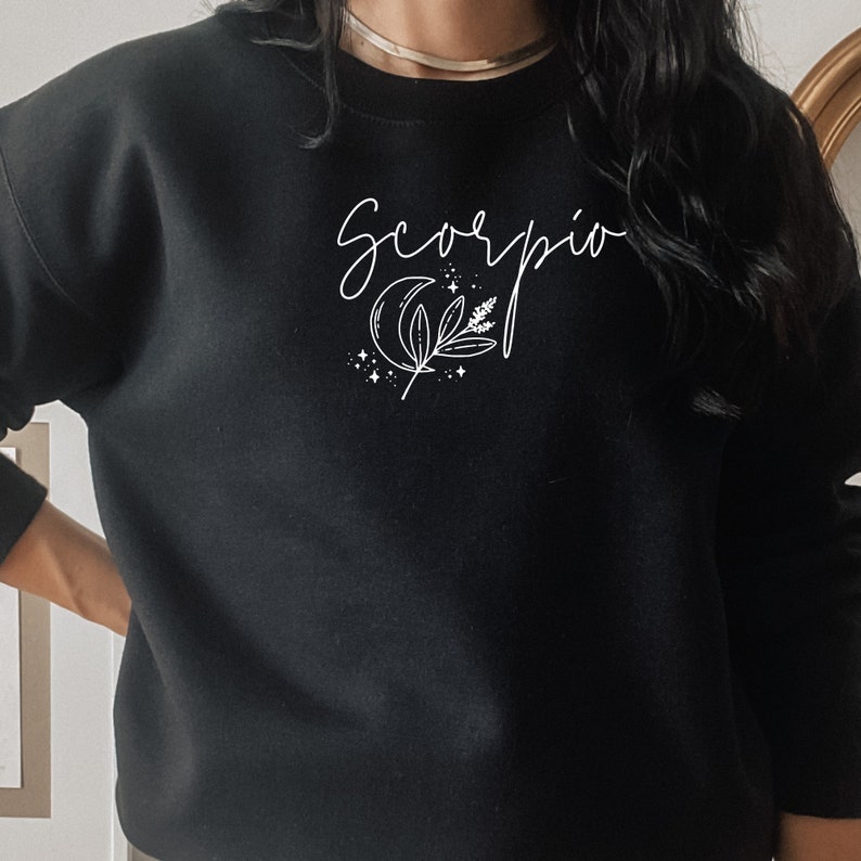 May include: Black crewneck sweatshirt with the word "Scorpio" in a cursive font. A white crescent moon, sprig, and stars graphic is below the word. The sweatshirt is a solid black color.