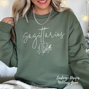 Puede incluir: Una sudadera verde con el texto "Sagittarius" en escritura blanca, una luna creciente, una rama de hojas y estrellas. La sudadera est&aacute; sobre una superficie blanca.