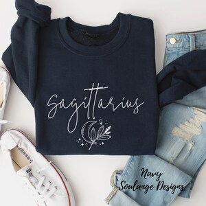Puede incluir: Sudadera azul marino con un gr&aacute;fico blanco del signo zodiacal Sagitario, una luna creciente y una rama de hojas. El texto "Sagitario" est&aacute; escrito en una fuente cursiva. La sudadera est&aacute; colocada sobre una superficie blanca con un par de zapatillas blancas y unos vaqueros azules.