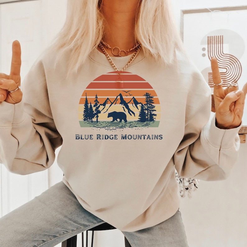 May include: A beige sweatshirt with a vintage-style graphic of mountains, a bear, and a sunset. The text "BLUE RIDGE MOUNTAINS" is printed below the graphic.