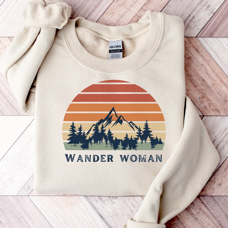K&ouml;nnte beinhalten: Ein beiger Pullover mit einem Vintage-Sonnenuntergang-Motiv mit einer Bergkette und B&auml;umen. Der Text "WANDER WOMAN" ist unter dem Motiv gedruckt.