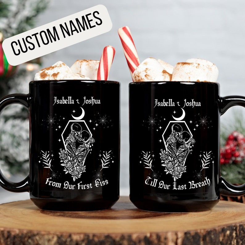 K&ouml;nnte beinhalten: Zwei schwarze Kaffeetassen mit wei&szlig;en Designs. Die Tassen zeigen ein Paar Skelette, die sich mit einem Halbmond und Sternen im Hintergrund umarmen. Eine Tasse tr&auml;gt die Aufschrift "Isabella & Joshua From Our First Kiss" und die andere "Isabella & Joshua Till Our Last Breath".