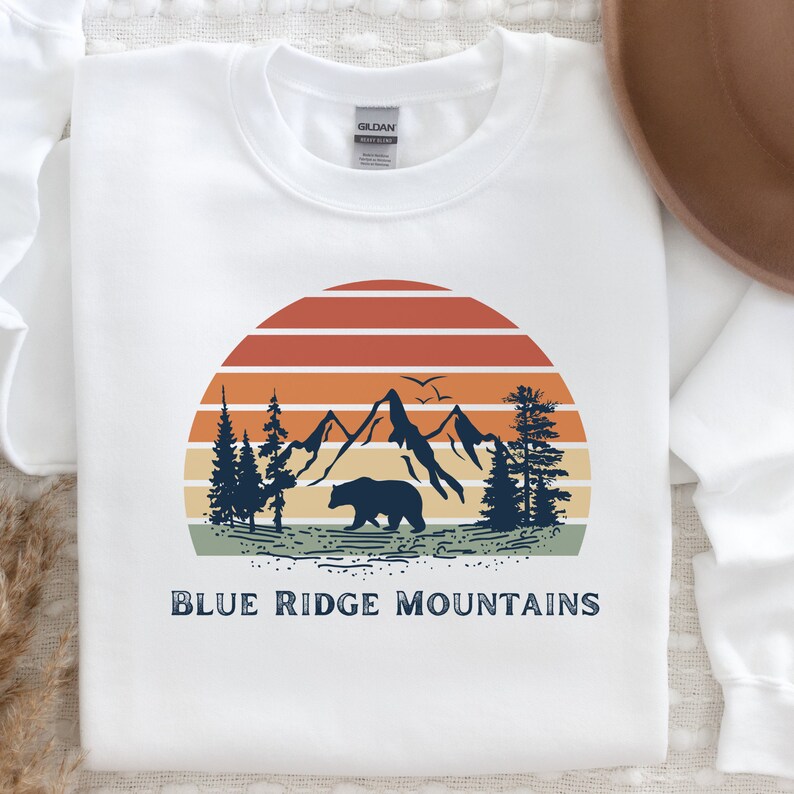 May include: White sweatshirt with a vintage-style graphic of a sunset over mountains, trees, and a bear. The text "BLUE RIDGE MOUNTAINS" is printed below the graphic.