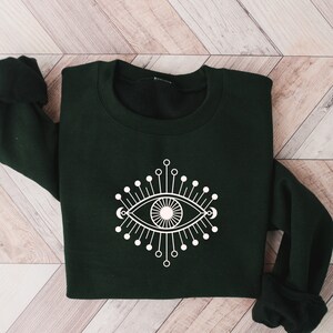 May include: A dark green sweatshirt with a white graphic of a stylized eye with radiating lines.