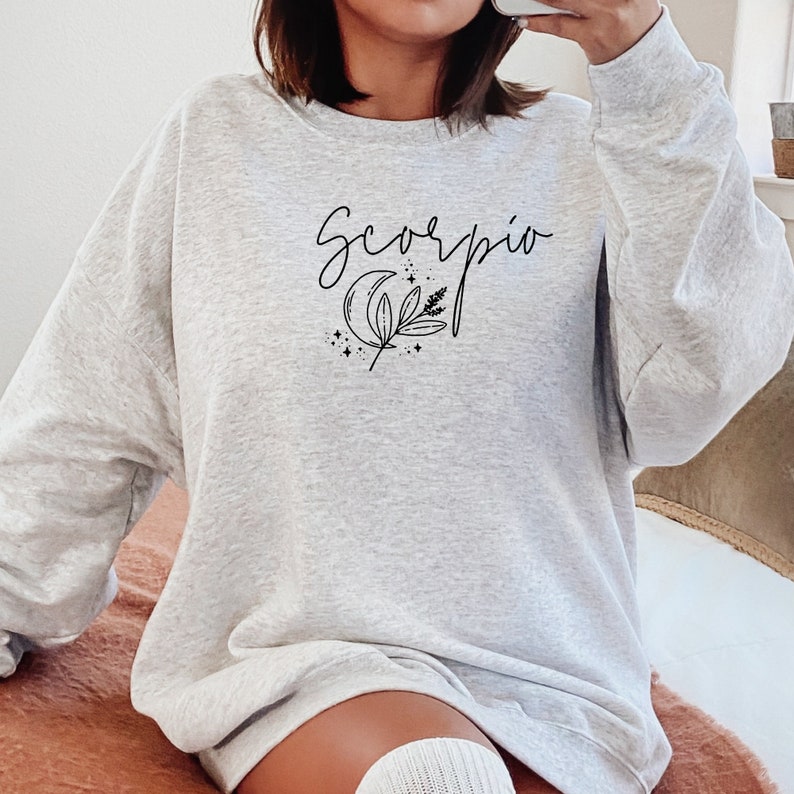 May include: Light gray sweatshirt with the word "Scorpio" in cursive script, along with a crescent moon and botanical illustration. The design is in black. The sweatshirt is a casual style.