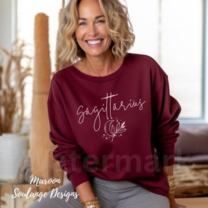Puede incluir: Una sudadera color granate con el texto "Sagitario" en letras blancas cursivas y una luna creciente blanca y un dise&ntilde;o floral.