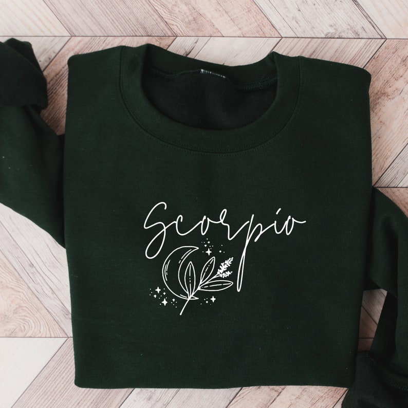 May include: A dark green sweatshirt with the word "Scorpio" in a white script font. Below the word is a white graphic of a crescent moon, leaves, and stars. The sweatshirt is folded on a light wood surface.