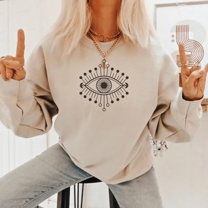 May include: A beige sweatshirt with a black and white graphic of an eye with rays emanating from it.