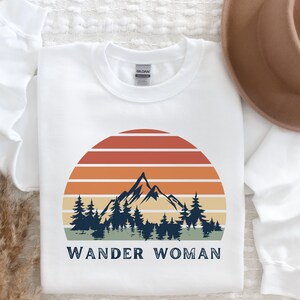 K&ouml;nnte beinhalten: Wei&szlig;er Pullover mit einem Retro-Sonnenuntergang-Motiv mit einer Bergkette und Kiefern. Der Text "WANDER WOMAN" ist unter dem Motiv gedruckt.