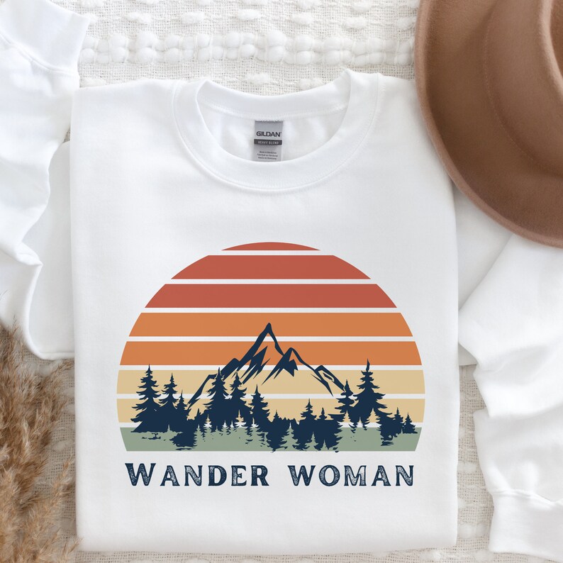 K&ouml;nnte beinhalten: Wei&szlig;er Pullover mit einem Retro-Sonnenuntergang-Motiv mit einer Bergkette und Kiefern. Der Text "WANDER WOMAN" ist unter dem Motiv gedruckt.