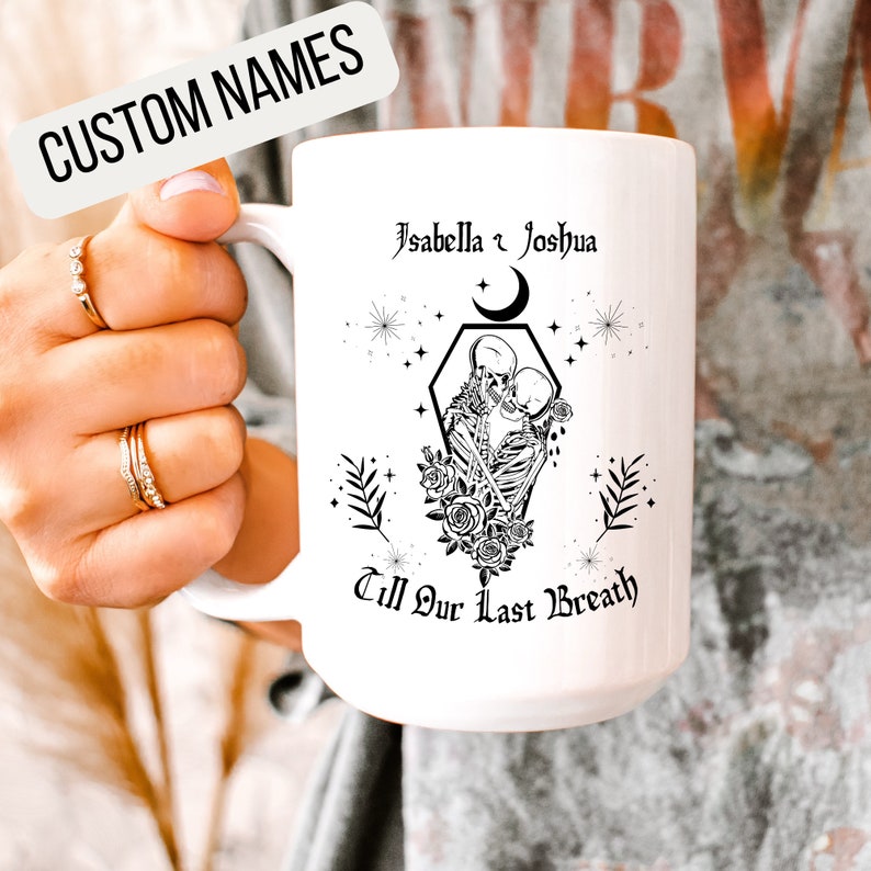 K&ouml;nnte beinhalten: Eine wei&szlig;e Keramiktasse mit einer schwarz-wei&szlig;en Grafik von zwei Skeletten, die sich umarmen, mit einer Mondsichel und Sternen. Der Text "Isabella & Joshua Till Our Last Breath" ist auf die Tasse gedruckt.