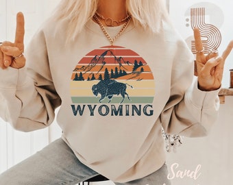 May include: A beige sweatshirt with a vintage-style graphic of a mountain range, a bison, and the word "WYOMING" in a sunset color palette.