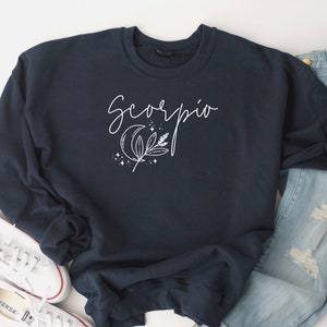 May include: Navy blue sweatshirt with the word "Scorpio" in cursive script. A white graphic of a crescent moon, stars, and foliage is below the word. The sweatshirt is paired with blue jeans and white sneakers.