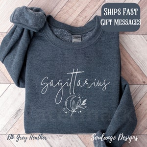 Puede incluir: Sudadera de color gris oscuro jaspeado con texto blanco que dice "Sagitario" y un dise&ntilde;o estilizado de luna, estrellas y hojas.