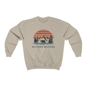 May include: A beige sweatshirt with a vintage-style graphic of a bear walking in front of mountains with a sunset behind it. The text "BLUE RIDGE MOUNTAINS" is printed below the graphic.