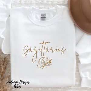 Puede incluir: Sudadera blanca con un dise&ntilde;o gr&aacute;fico marr&oacute;n que dice "Sagitario" con una luna creciente y una rama de hojas. El texto "Soulance Designs White" est&aacute; en la parte inferior de la sudadera.