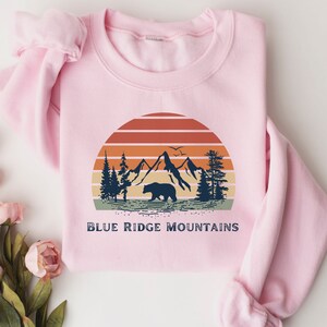 May include: Pink sweatshirt with a vintage-style graphic of mountains, a bear, and a sunset. The text "BLUE RIDGE MOUNTAINS" is printed below the graphic.