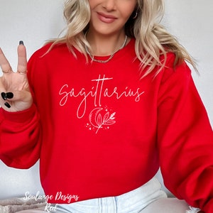 Puede incluir: Sudadera roja con texto blanco que dice "Sagitario" y una luna creciente blanca y un dise&ntilde;o floral.