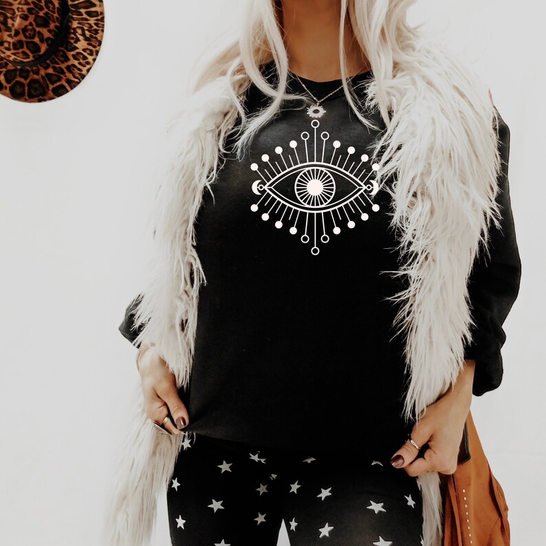 May include: Black sweatshirt with a white graphic of a stylized eye with radiating lines and circles. The sweatshirt is worn over a white faux fur vest and black pants with a white star pattern.