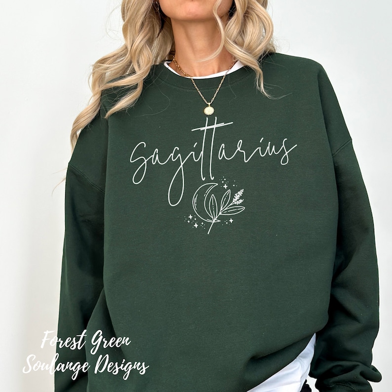 Puede incluir: Sudadera verde bosque con texto blanco que dice "Sagitario" y una luna creciente con un dise&ntilde;o de hoja.