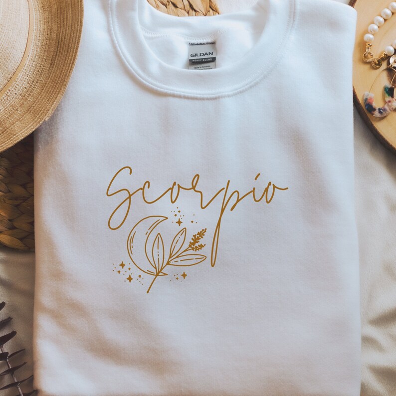 May include: White sweatshirt with the word "Scorpio" in gold script, along with a gold crescent moon, sprig, and stars design. The Gildan brand label is visible on the collar. The sweatshirt is a casual, comfortable garment.