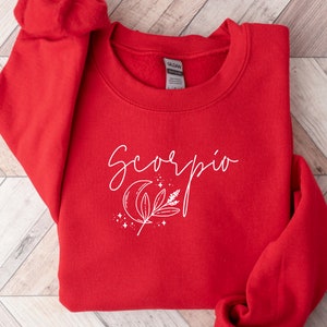 May include: A red crewneck sweatshirt with the word "Scorpio" in white script. Below the word is a white graphic of a crescent moon, leaves, and stars. The sweatshirt is made of a soft material.