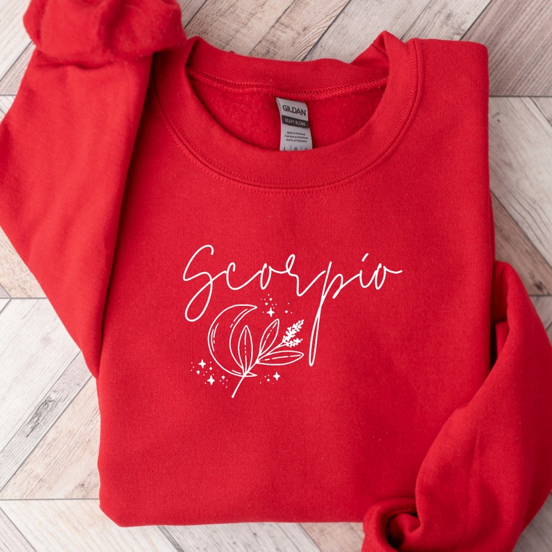 May include: A red crewneck sweatshirt with the word "Scorpio" in white script. Below the word is a white graphic of a crescent moon, leaves, and stars. The sweatshirt is made of a soft material.