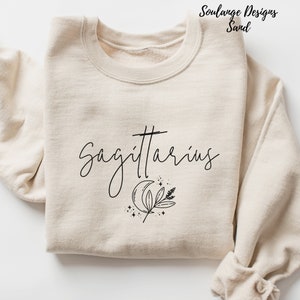 Puede incluir: Una sudadera beige con el texto "Sagittarius" en letras cursivas negras. Tambi&eacute;n se imprime una luna creciente, una hoja y estrellas en la sudadera.