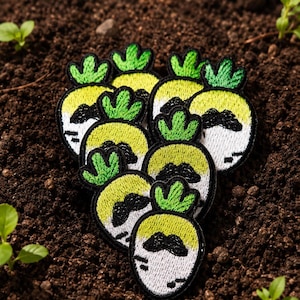 May include: A collection of embroidered patches shaped like cartoon radishes with green tops, yellow bodies, and black mustaches. The patches are arranged on a bed of dark brown soil, with small green plants visible.