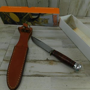 May include: A fixed-blade knife with a stacked leather handle and a silver-colored pommel, accompanied by a tan leather sheath with white stitching and a snap closure. The knife is displayed next to its orange and white cardboard packaging box.