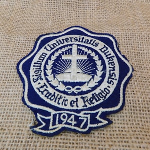 May include: A blue and white embroidered patch with the text "Sigillum Universitatis Dukensis Traditio et Religio 1947". The patch features a cross and a wreath design.