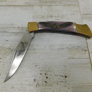 May include: A folding pocket knife with a polished stainless steel blade featuring an etched eagle design. The handle has brass bolsters and a decorative inlay depicting a flying eagle against a muted, light-colored background with subtle textures.