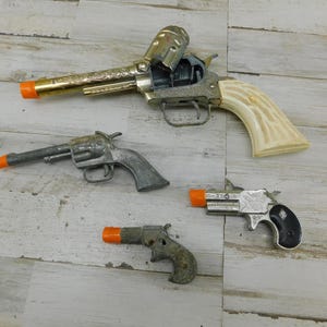 May include: Four toy cap guns are displayed on a light wood surface. The largest gun has a gold barrel and a cream-colored handle. The other three guns are smaller, with silver and black finishes and orange tips.