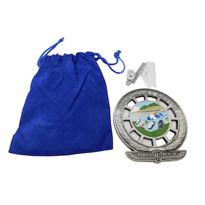 May include: A silver-colored medallion with the text "70th Concours d'Elegance" and "Pebble Beach" is displayed. The medallion features a colorful image of a vintage race car. A blue velvet drawstring bag and a clear plastic stand are also included.