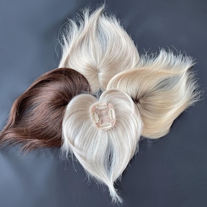 Op de afbeelding: Een assortiment haartoppers in verschillende tinten bruin, blond en wit. De haarstukken zijn gerangschikt op een donkergrijze ondergrond, met verschillende stijlen en kleuren. Eén topper heeft clips voor bevestiging.