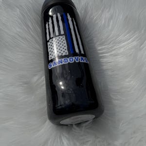 May include: Black water bottle with a blue and white American flag design and the name "Sandoval" in blue. The bottle has a blue lid and a black body.